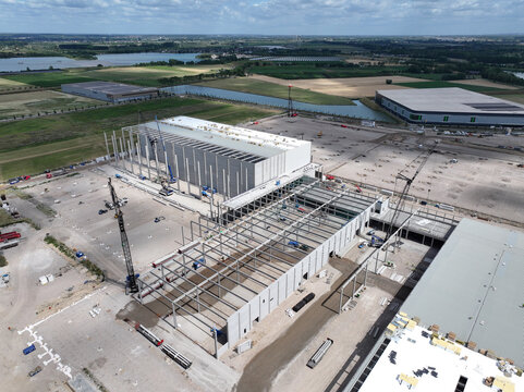 Aerial view of a large industrial warehouse construction site featuring steel framework, cranes, and concrete foundations surrounded by open fields and canals in the Netherlands.