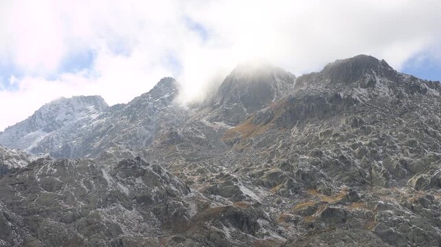 Rocky alpine peaks with first snow near Airolo, Switzerland. Static shot of clouds drifting over a mountain ridge in the Swiss Alps, autumn scree slopes and misty summits in daylight