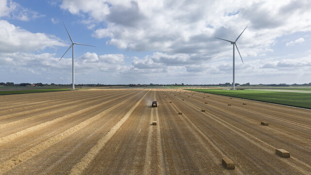 Aerial view of a tractor harvesting hay in a vast field with scattered hay bales and two large wind turbines under a blue sky in Middenmeer, North Holland, Netherlands.