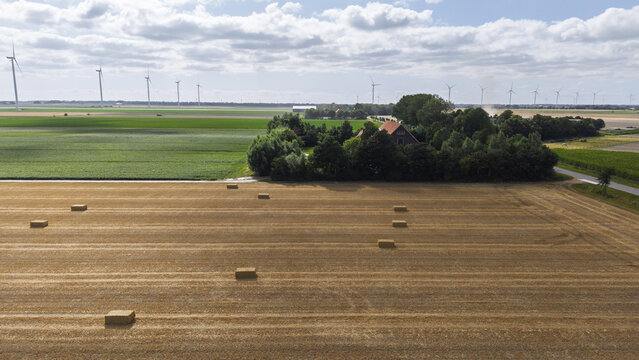 Aerial view of agricultural fields with hay bales and a farmhouse surrounded by trees under a cloudy sky in Middenmeer, North Holland, Netherlands.