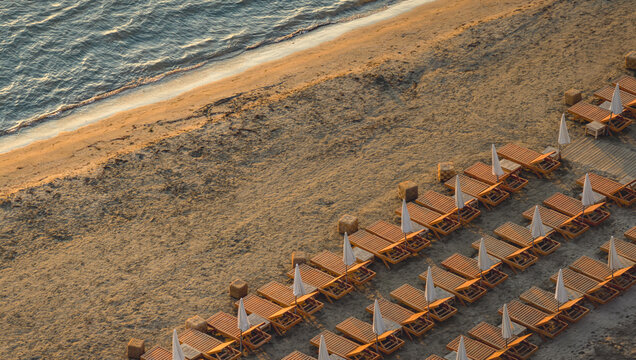 Aerial view of neatly arranged wooden sun loungers and white umbrellas on a sandy beach at golden hour in Le Grau-du-Roi, Occitanie, France.