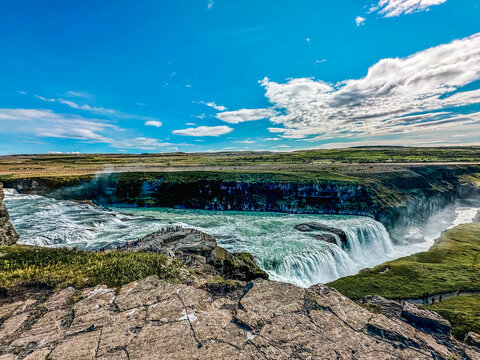 View of Gullfoss waterfall cascading over rocky cliffs with lush green landscape and tourists on the viewing platform under a blue sky in Iceland.
