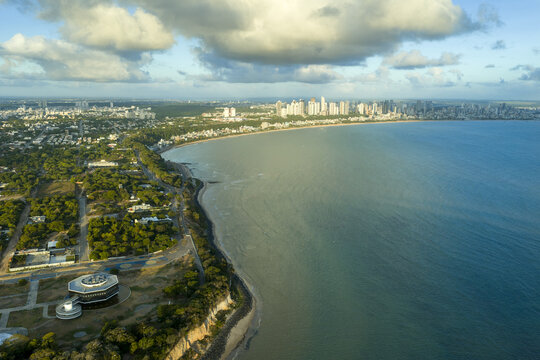 Aerial view of the coastline at Ponta do Seixas, featuring the Estacao Cabo Branco, lush vegetation, and the skyline of Joao Pessoa along the Atlantic Ocean in Joao Pessoa, Paraiba, Brazil.