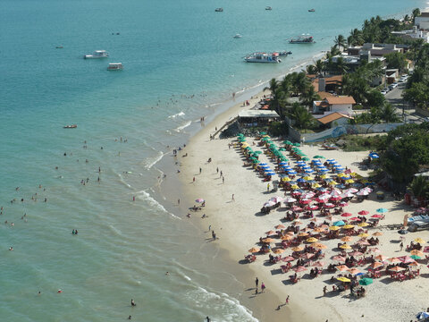 Aerial view of Seixas Beach with colorful umbrellas, people relaxing on the sand, boats in the turquoise water, and coastal buildings in Joao Pessoa, Paraiba, Brazil.