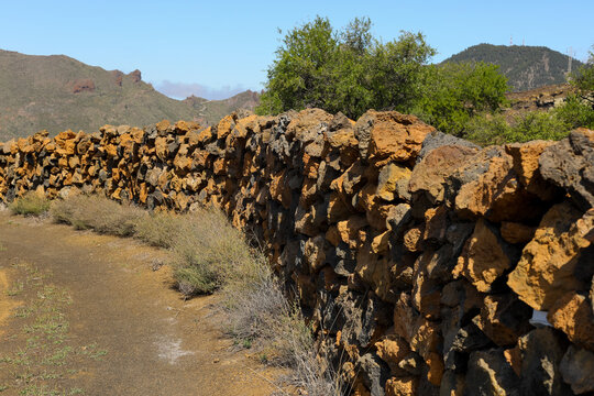 View of a dry stone wall made of volcanic rock curving along a dirt path in a rugged landscape with green bushes and mountains in Santa Cruz de Tenerife, Canary Islands, Spain.