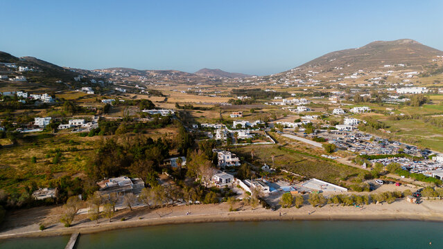 Aerial view of the coastal landscape featuring white buildings, sandy beaches, and rolling hills under a clear blue sky in Paros, Greece.