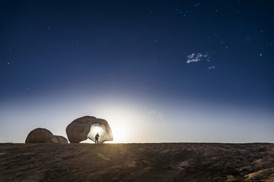 View of a person standing under the unique rock formation of Lajedo de Pai Mateus at night, illuminated by light against a starry sky in the rocky landscape of Cabaceiras, Paraiba, Brazil.