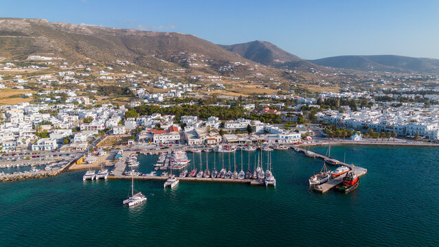 Aerial view of the harbor in Paros with docked sailboats and catamarans, surrounded by white buildings and coastal hills in Paros, Greece.