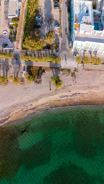 Aerial view of a sandy beach meeting the clear turquoise sea, with a coastal road, parked cars, and white buildings in Paros, Greece.