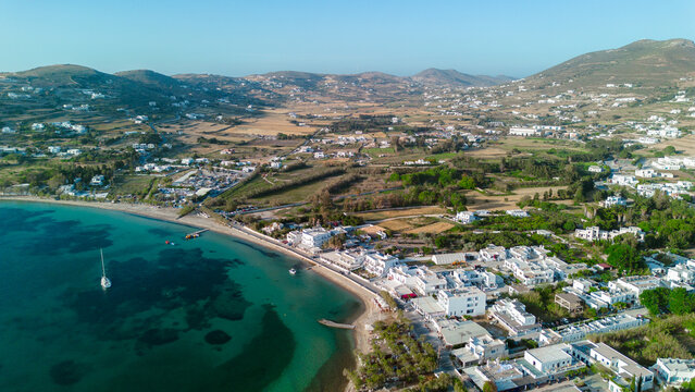 Aerial view of the coastal town of Paros, featuring white buildings, a sandy beach, turquoise waters, and rolling hills in the background in Paros, Greece.