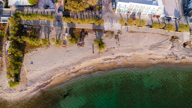 Aerial view of a sandy beach with turquoise water, lined with trees and a coastal road with buildings in Paros, Greece.