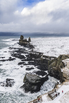 Aerial view of the dramatic Londrangar basalt volcanic plugs rising from the snow-covered coastline with crashing waves and dark volcanic rocks in Hellnar, Snaefellsbaer, Iceland.