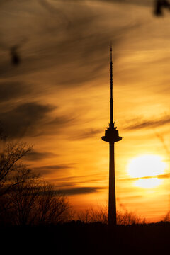 View of the Vilnius TV Tower silhouetted against a vibrant golden sunset sky with bare tree branches in the foreground in Vilnius, Vilnius County, Lithuania.