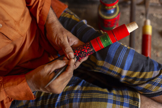 View of a craftsman meticulously painting intricate floral patterns onto a wooden object with vibrant red, green, and black glazes in Kashmore, Sindh, Pakistan.