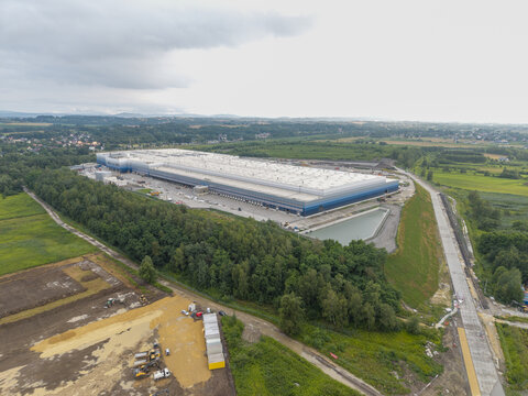 Aerial view of the Lidl logistics center under construction, featuring a large warehouse building, surrounding green fields, trees, and a paved road in Skawina, Lesser Poland Voivode, Poland.