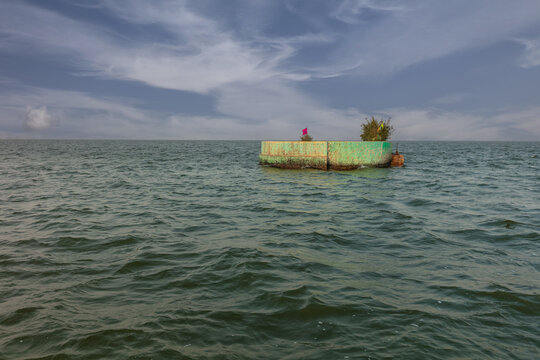 View of the small circular concrete structure in Keenjhar Lake with sparse vegetation and a small flag under a cloudy sky in Thatta, Sindh, Pakistan.