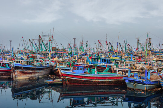 View of colorful wooden fishing boats docked in the harbor with calm water reflections under a cloudy sky in Karachi, Sindh, Pakistan.