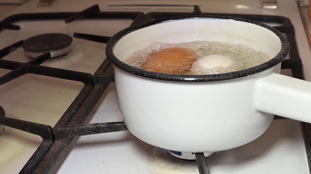 Close-up boiling eggs in enameled ladle on gas stove, brown and white chicken eggs simmering in bubbling water, steam rising, white