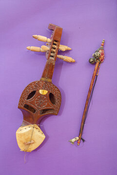 View of a traditional wooden Sarangi musical instrument with its bow, featuring intricate carvings and a skin-covered resonator, placed against a solid purple background in Hyderabad, Sindh, Pakistan.