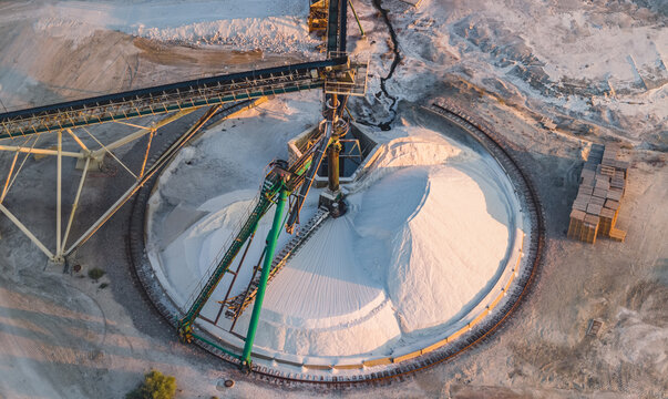 Aerial view of an automated conveyor system stacking harvested sea salt into a circular stockpile at an industrial salt production facility during golden hour, France.