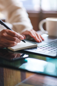 Vertical closeup image of a woman writing on a notebook while working on laptop computer