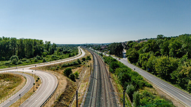 Aerial view of a railway line running parallel to a road, surrounded by lush green trees and vegetation under a clear blue sky in Sremski Karlovci, Vojvodina, Serbia.