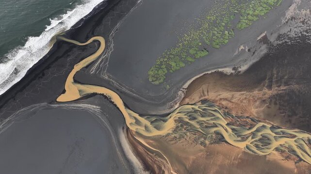 Aerial view of braided river delta with sediment-rich water flowing into the sea across a black sand beach with patches of green moss in Iceland.