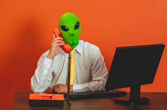 A man in an alien mask talks on a retro telephone at the office