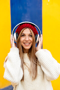 Smiling woman in red beanie listening to music on headphones against yellow background
