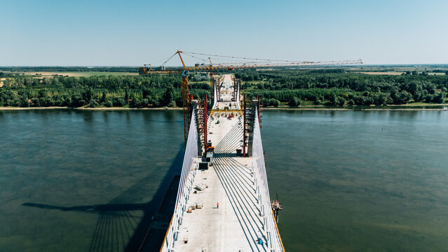 Aerial view of the construction of a cable-stayed bridge over a wide river with a crane and workers on the concrete deck in Sremski Karlovci, Vojvodina, Serbia.
