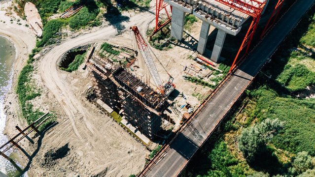 Aerial view of a bridge construction site featuring a tall crane, concrete support pillars, and scaffolding near the riverbank in Sremski Karlovci, Vojvodina, Serbia.