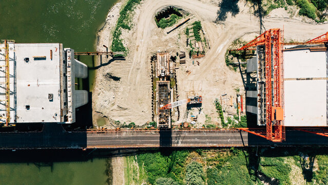 Aerial view of a bridge construction site with concrete piers, orange cranes, and heavy machinery on the riverbank in Sremski Karlovci, Vojvodina, Serbia.