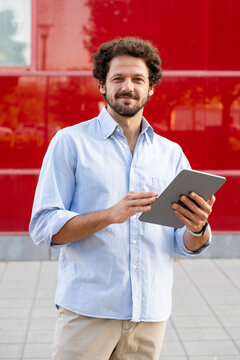 Smiling businessman holds a digital tablet outdoors by a red wall