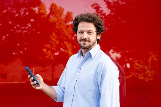 Smiling entrepreneur holds a smartphone outdoors by a red wall