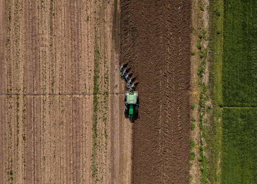 Aerial view of a tractor plowing a field during the spring sowing season, showing the contrast between tilled dark soil and adjacent green crops in Sesto Ulteriano, Lombardy, Italy.