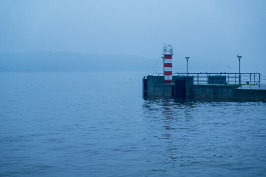 View of a red and white striped lighthouse on a concrete pier surrounded by calm, misty water in Klaipeda, Klaipeda County, Lithuania.
