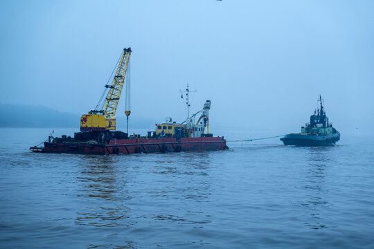 View of a tugboat towing a barge equipped with a yellow crane across the calm waters of the harbor under a misty sky in Klaipeda, Klaipeda County, Lithuania.