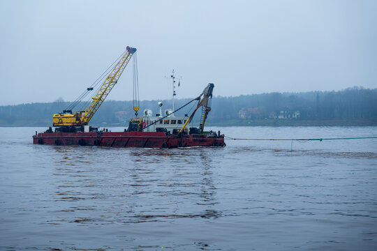 View of a red dredging vessel with a yellow crane operating on the river under a cloudy sky in Klaipeda, Klaipeda County, Lithuania.