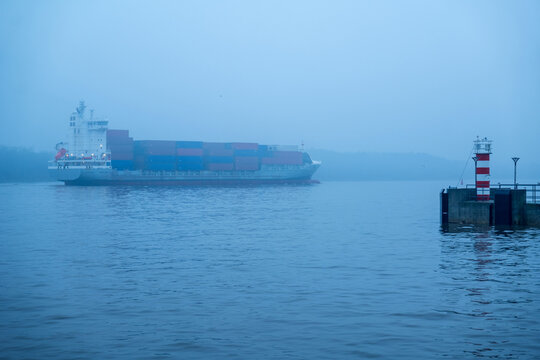 View of a large container ship sailing through the misty waters near the red and white lighthouse in the port of Klaipeda, Klaipeda County, Lithuania.