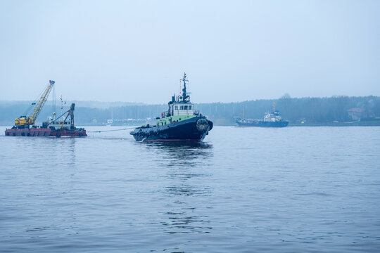 View of a tugboat navigating the harbor waters with a crane barge nearby under a misty sky in Klaipeda, Klaipeda County, Lithuania.