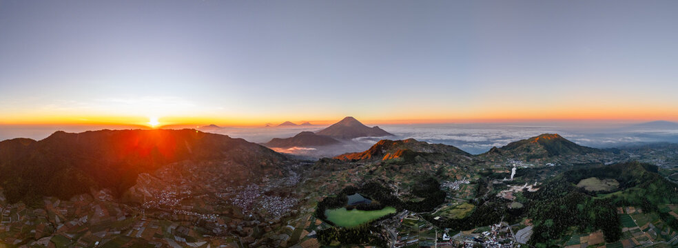 Aerial view of the Dieng Plateau at sunrise, featuring the vibrant volcanic lake, surrounding mountains, and terraced agricultural fields in Kejajar, Central Java, Indonesia.