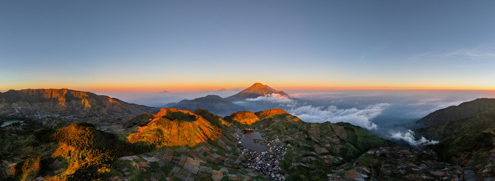 Aerial view of the volcanic landscape of Dieng Plateau at sunset, featuring terraced agricultural fields, a mountain lake, and distant peaks in Kejajar, Central Java, Indonesia.