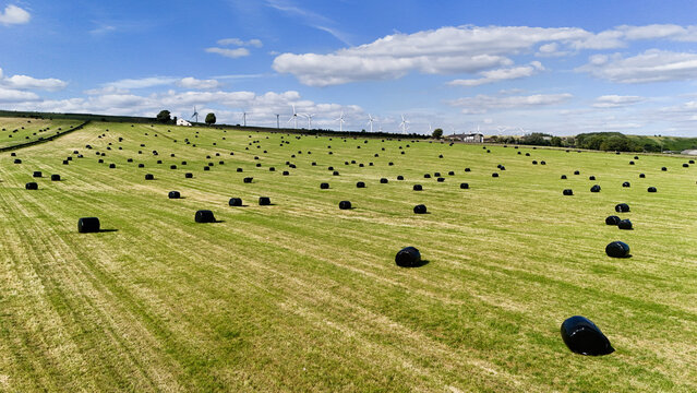 Aerial view of a green field dotted with black plastic-wrapped hay bales under a blue sky with wind turbines in the background in Millhouse Green, England, United Kingdom.