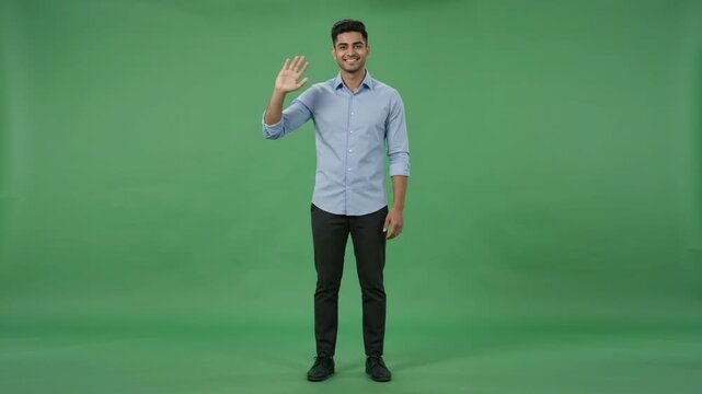 Friendly young man in blue shirt waving hello against a green background, full body shot.