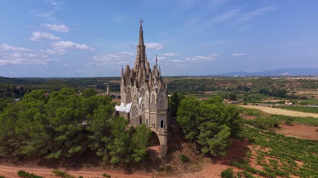 Aerial view of the Santuari de la Mare de Deu de Montserrat de Montferri with catenary arch spires above pine trees, surrounded by vineyards and farmland in Tarragona, Catalonia