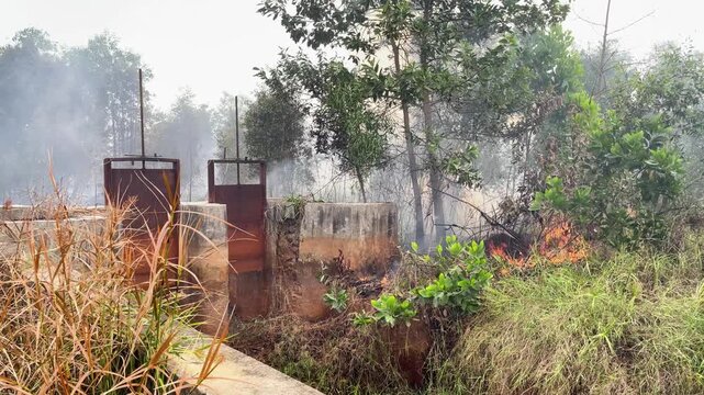 Aerial view of a forest fire burning near a concrete sluice gate with smoke rising through trees and dry vegetation in Banjarbaru, South Kalimantan, Indonesia.