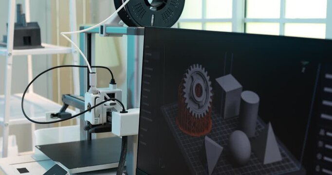 3d printer in a workshop beside a monitor a digital 3d model of gears and shapes, with cables and equipment in a modern maker space.