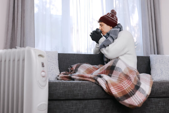 Freezing man at home in winter clothes. Struggling with cold indoor heating, he drinks hot tea by a heater, facing the energy crisis.