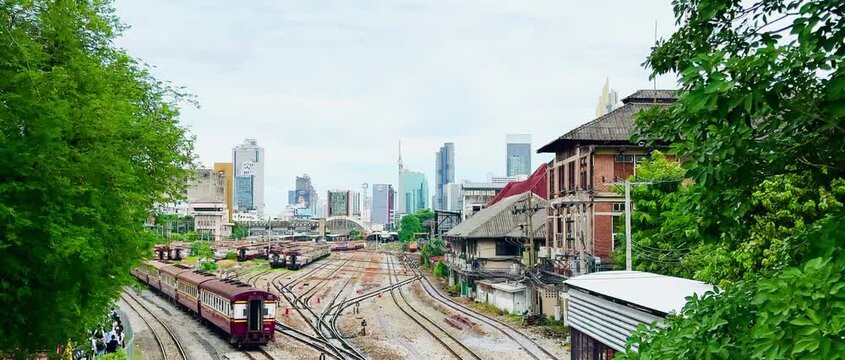 High-angle view of vintage passenger trains navigating multiple railway tracks at Hua Lamphong Station yard with the modern Bangkok city skyline and overcast sky in the background.
