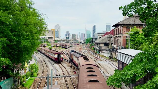 High-angle view of vintage passenger trains navigating multiple railway tracks at Hua Lamphong Station yard with the modern Bangkok city skyline and overcast sky in the background.